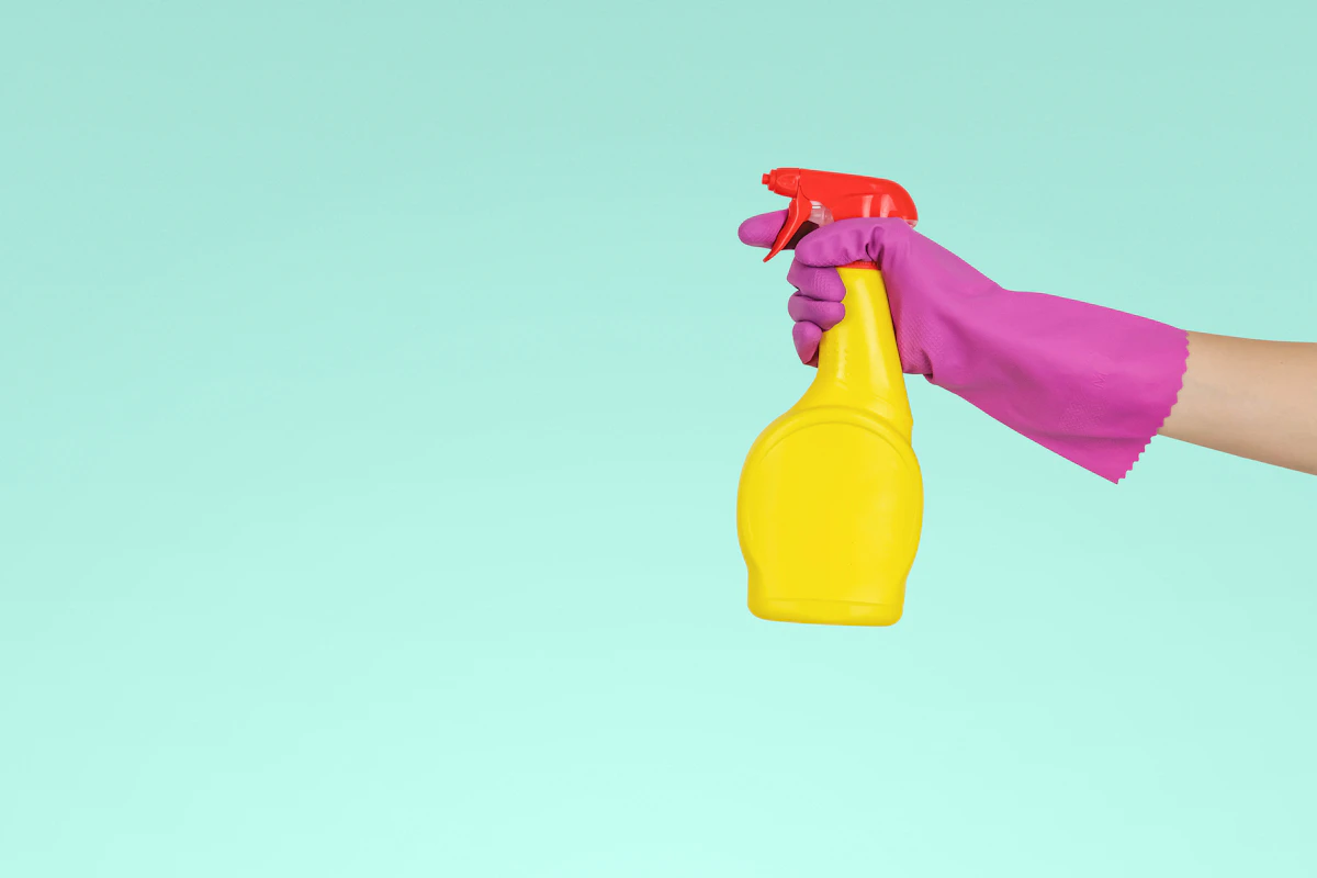Purple-gloved hand holding a yellow cleaning spray bottle against a mint background